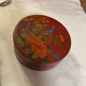May include: A round, red decorative box with a lid. The lid features a painted design of two peacocks, orange flowers, and green foliage. The box is made of a dark red material.