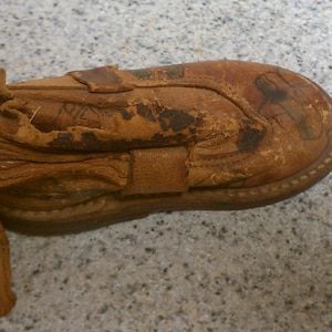 May include: A worn brown leather baby shoe with a strap and a frayed edge. The shoe has a worn leather sole and is decorated with faded markings.