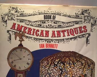 May include: Book cover with the title "Book of American Antiques" by Ian Bennett. The cover features a vintage clock with an eagle finial and a stained glass lampshade.