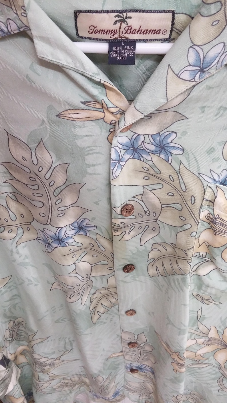 May include: A light green silk button-down shirt with a tropical print featuring palm trees, flowers, and leaves. The shirt has a Tommy Bahama label and is made in China.