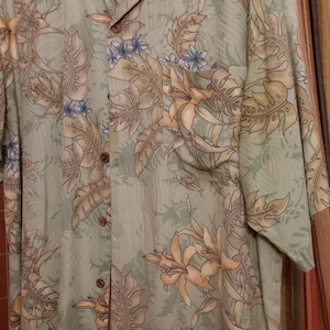 May include: A green, short-sleeved button-down shirt with a floral print. The shirt features a light green background with brown, yellow, and blue floral patterns. The shirt has a pocket on the left side and a Tommy Bahama label on the collar.