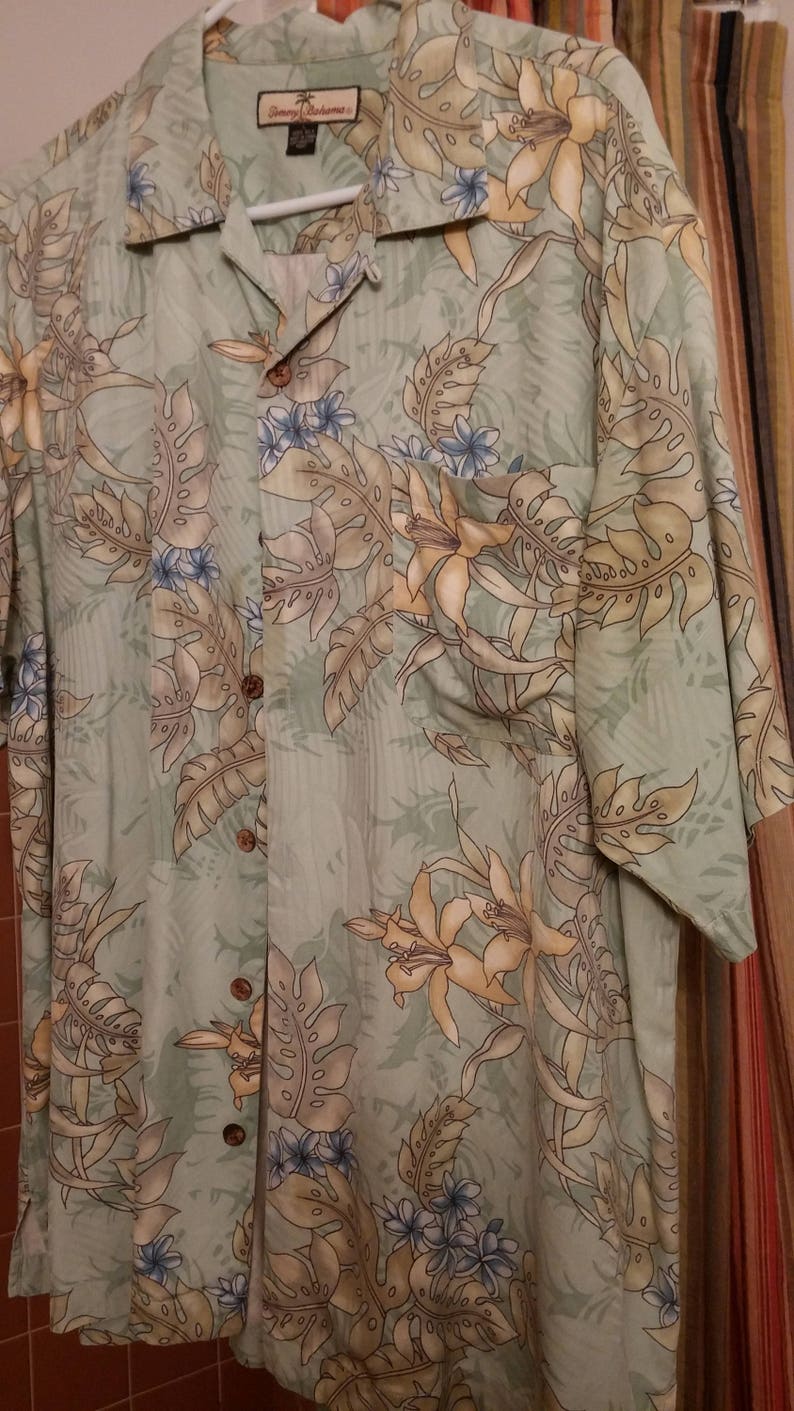 May include: A green, short-sleeved button-down shirt with a floral print. The shirt features a light green background with brown, yellow, and blue floral patterns. The shirt has a pocket on the left side and a Tommy Bahama label on the collar.