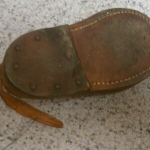 May include: The underside of a brown leather shoe with a stitched seam and a leather strap. The shoe has several metal rivets.