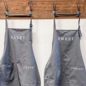 Two grey aprons, on says salty and the other says sweet, hanging on a wall