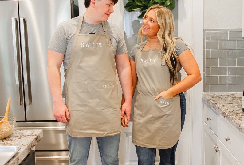 A man and woman standing in the kitchen wearing beige aprons and holding hands