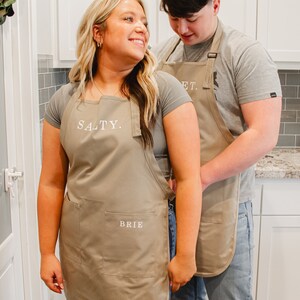 A man and woman standing in the kitchen wearing beige aprons. The man is tying the woman's apron.