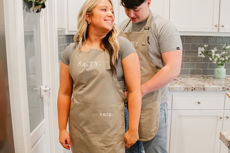 A man and woman standing in the kitchen wearing beige aprons. The man is tying the woman's apron.