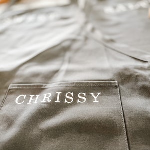 A grey apron with a pocket that has the name "Chrissy" printed on it