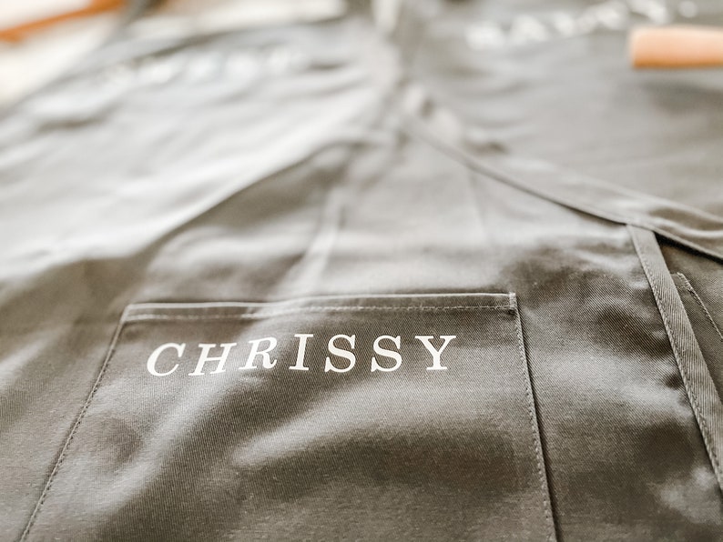 A grey apron with a pocket that has the name "Chrissy" printed on it
