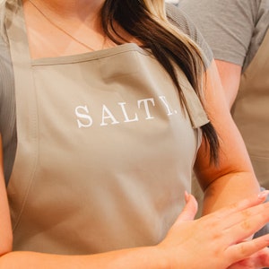 A woman with a beige apron that says salty on it with a man wearing a beige apron behind her