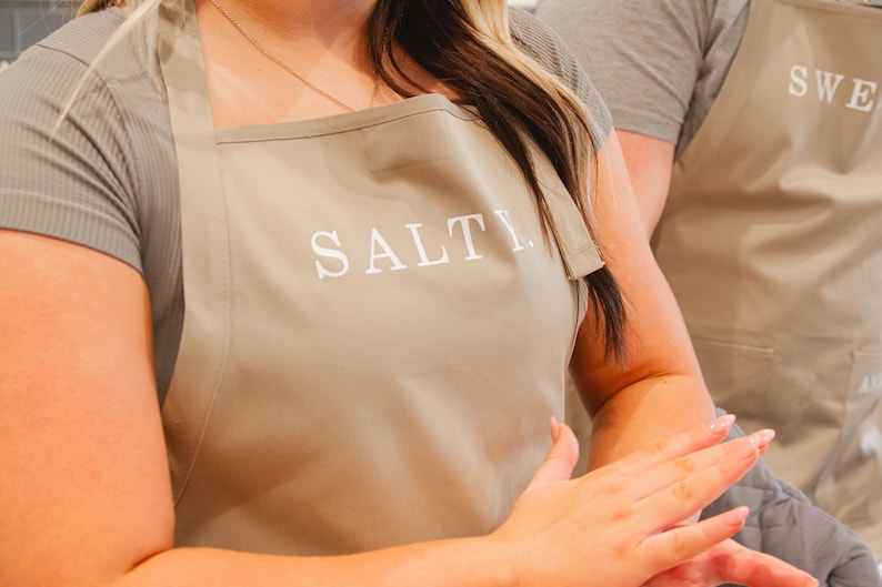 A woman with a beige apron that says salty on it with a man wearing a beige apron behind her