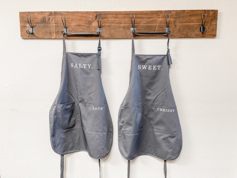 Two grey aprons, on says salty and the other says sweet, hanging on a wall