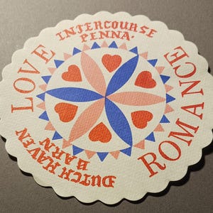Könnte beinhalten: Ein runder weißer Untersetzer mit einem gewellten Rand. Die Mitte zeigt ein blaues und rosafarbenes Sternburst-Design mit roten Herzen. Der Text "LOVE" befindet sich auf der linken Seite, "ROMANCE" auf der rechten Seite und "DUTCH HAVEN BARN INTERCOURSE PENNA." befindet sich oben herum.