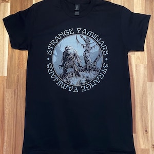 May include: Black t-shirt featuring a circular graphic with the text "STRANGE FAMILIARS." The graphic depicts a creature in a forest setting. The design is in shades of gray and white, with the text in a stylized font.