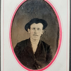 May include: A black and white portrait of a man wearing a black hat and a dark jacket. The photo is in an oval frame with a pink border.
