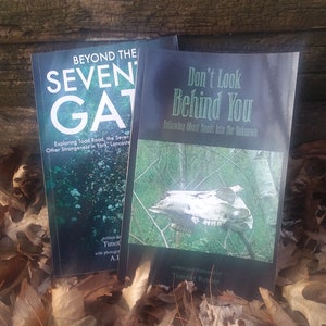 Può includere: Due libri tascabili con copertine nere e testo bianco. Il libro a sinistra è intitolato "Beyond the Seven Gates" e il libro a destra è intitolato "Don't Look Behind You: Following Ghost Roads into the Unknown".