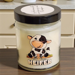May include: A glass jar candle with a black lid. The candle has a label with a cartoon cow and the words "NOT TODAY HEIFER". The candle is on a wooden surface.