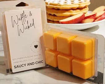 May include: A clear plastic package contains six orange wax melt cubes. A second package displays a white label with the text "Waffle & Wicked" and "Saucy and Chic". A stack of waffles and sliced apples are in the background.