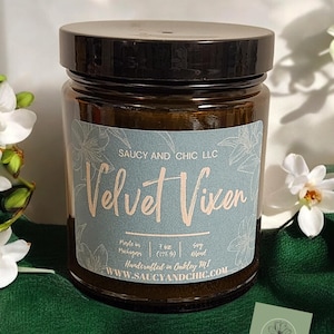 May include: A brown glass candle jar with a black lid. The label is light blue with a floral design and the text "SAUCY AND CHIC LLC Velvet Vixen Made in Michigan 7 oz (198 g) Soy Blend Handcrafted in Oakley MI WWW.SAUCYANDCHIC.COM".