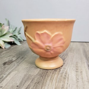 May include: A vintage peach-colored ceramic planter with a raised floral design. The planter has a wide, round base and a flared top. The flower design is in shades of pink and yellow.
