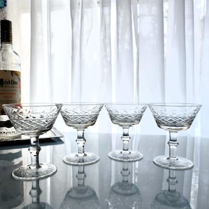 May include: Four clear, stemmed champagne coupe glasses with diamond-patterned bowls and etched floral designs. The glasses are arranged on a reflective surface, with a bottle of liquor visible in the background. The glasses are elegant and suitable for serving cocktails.