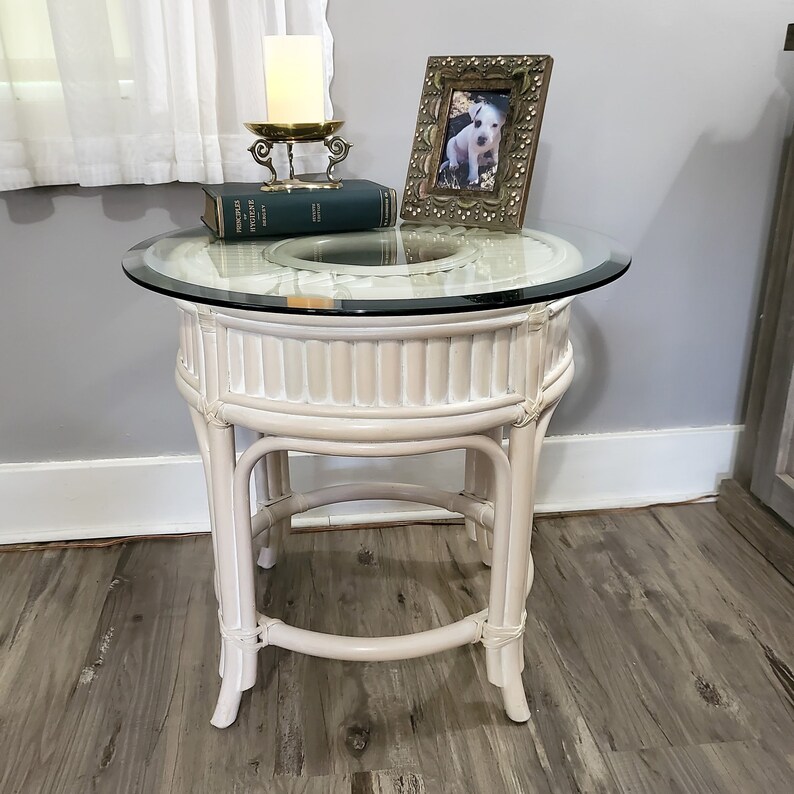 May include: White wicker end table with a glass top. The table has a round top and three legs. A book and a decorative candle holder are on top of the table.