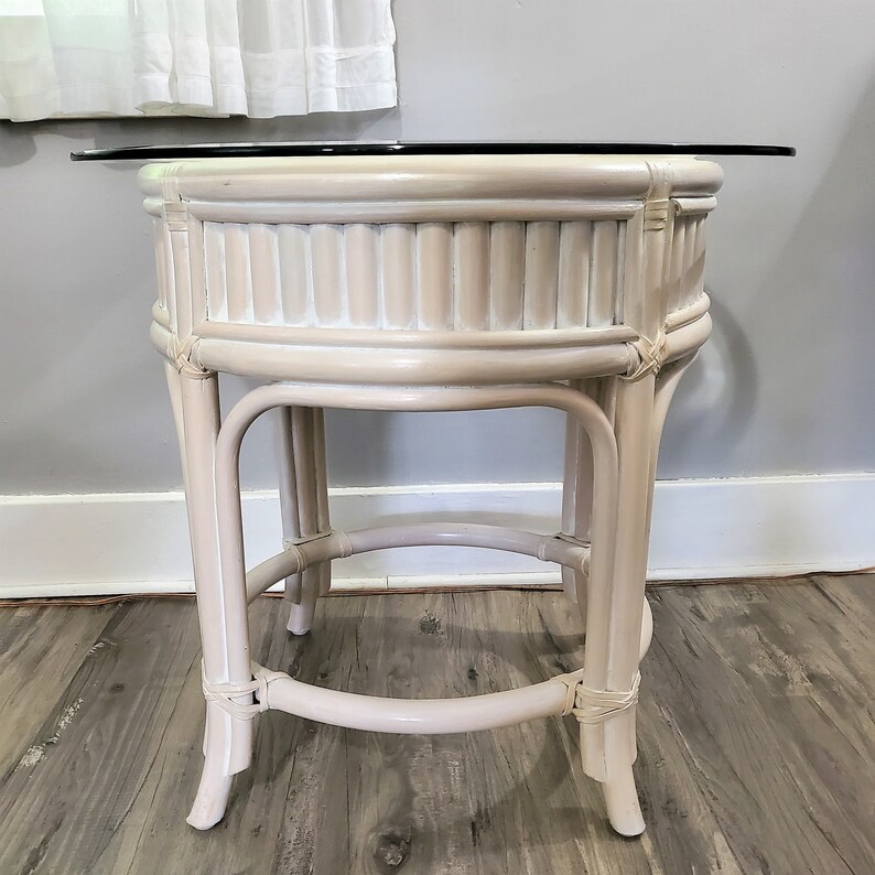 May include: A white wicker end table with a round glass top. The table has a woven design and a distressed white finish.