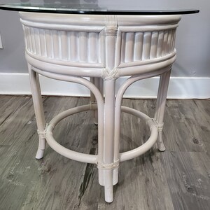 May include: White wicker end table with a round glass top. The table has a woven design and a simple, elegant style.