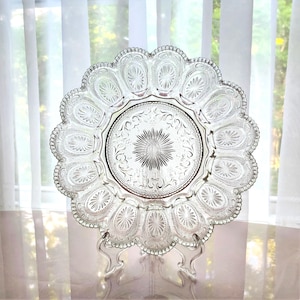 May include: Clear glass serving plate with a scalloped edge and intricate embossed floral design. The plate is displayed on a clear stand, showcasing its detailed pattern. The plate's center features a starburst design.