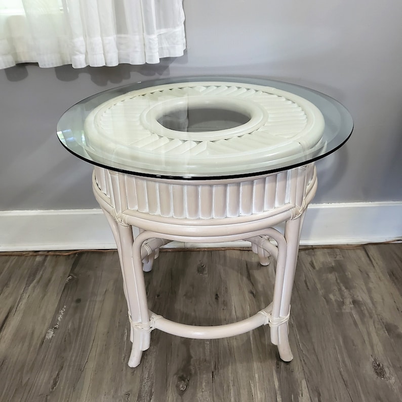 May include: A white wicker end table with a round glass top. The table has a decorative design on the top and a simple, sturdy base.