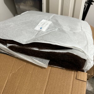 May include: A brown, rectangular cushion wrapped in white tissue paper. The cushion is sitting on top of a brown cardboard box.