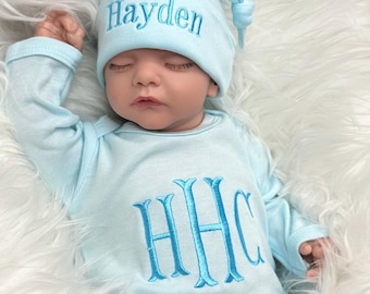 May include: A light blue baby onesie with long sleeves and a matching hat. The onesie has the name "Hayden" embroidered on the hat and the monogram "HHC" embroidered on the front. The baby is sleeping in the onesie and hat.