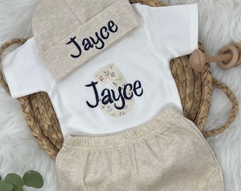 May include: A white baby onesie with a nautical themed 'J' and the name 'Jayce' embroidered on it. A beige knit hat with the name 'Jayce' embroidered on it. Beige knit pants.