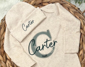 Può includere: Un body per neonato beige a maniche lunghe e chiusura a pressione sul fondo. Il body presenta un design ricamato verde e grigio con il nome "Carter" su di esso. È incluso anche un berretto beige abbinato con il nome "Carter" ricamato su di esso.