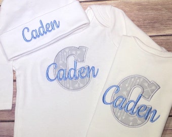 May include: A white baby outfit set with a long-sleeved bodysuit, a short-sleeved onesie, and a beanie hat. All three pieces feature the name "Caden" embroidered in blue with a gray and white patterned letter C.