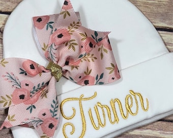 May include: White baby hat with a pink floral bow and the name "Turner" embroidered in gold thread.