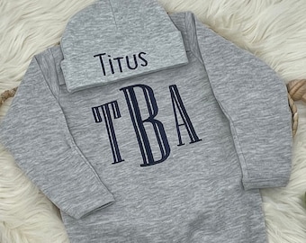 May include: A gray baby onesie and matching beanie set. The onesie features the embroidered letters "TBA" in navy blue, and the beanie is embroidered with the name "Titus". The set is made of a soft, comfortable material.