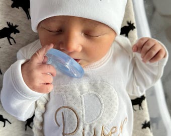 May include: A white baby onesie with a large embroidered letter 'R' and the name 'River' in gold thread. The baby is wearing a white knit hat with the name 'River' embroidered in gold thread.