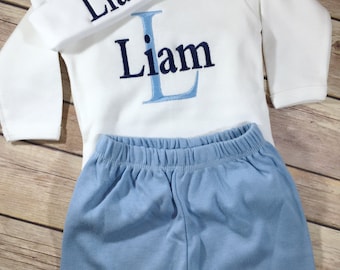 May include: A baby outfit set with a white hat, a white long-sleeved bodysuit, and blue pants. The hat and bodysuit are embroidered with the name "Liam" in navy blue thread. The bodysuit also has a large blue "L" embroidered on the front.