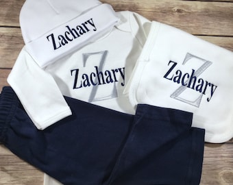 May include: A white baby outfit set with the name Zachary embroidered on each piece. The set includes a hat, a long-sleeved onesie, a bib, and a pair of navy blue pants.