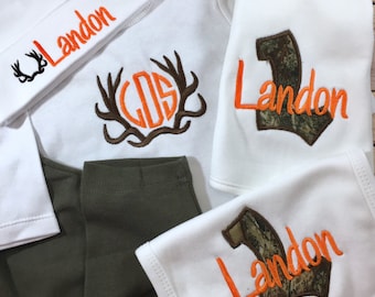 May include: A baby outfit set with a white hat, bodysuit, and bib. The hat and bodysuit have the name "Landon" embroidered in orange. The bodysuit has a monogram with antlers and the bib has a camouflage "L" with the name "Landon" embroidered in orange.