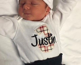 May include: A white baby onesie with a red and white checked number "3" and the name "Justin" embroidered in black. The onesie is worn by a baby wearing a white knitted hat with the name "Justin" embroidered in black. The baby is wearing black trousers.
