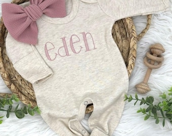 Op de afbeelding: Een beige babyromper met de naam "eden" in roze geborduurd. Een grote roze strik en een houten rammelaar staan ook in de afbeelding. De romper ligt in een gevlochten mand.