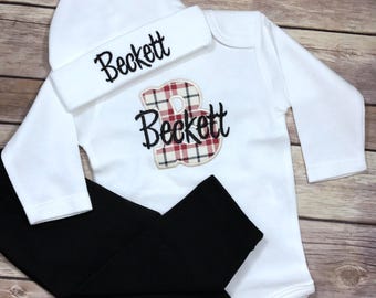 May include: A white baby outfit set with a long-sleeved onesie, black trousers, and a white beanie. The onesie has a red and white plaid letter "B" with the name "Beckett" embroidered on it. The beanie also has the name "Beckett" embroidered on it.