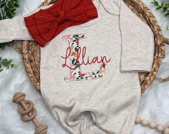May include: A beige baby onesie with the name "Lillian" embroidered in red script. The letter "L" is decorated with a floral pattern. A red textured bow headband rests above the onesie.