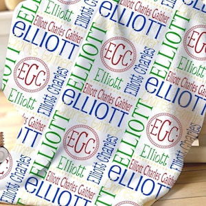 May include: A white blanket featuring a repeating pattern of the name Elliott in blue and green, the name Elliott Charles Gaither in red, and a circular monogram logo with the letters EGC in red, all printed across the fabric surface.