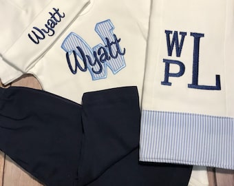 May include: A baby boy's outfit set in white and navy blue. The set includes a hat, a bodysuit, pants, and a blanket. The bodysuit and blanket are embroidered with the name "Wyatt" and the letter "W".