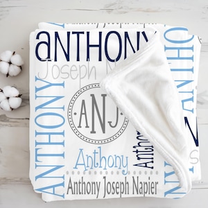 May include: A white fleece blanket featuring the name Anthony in various shades of blue and grey, along with the full name Anthony Joseph Napier and a circular monogram with the letters ANJ in the center, displayed in a folded arrangement.
