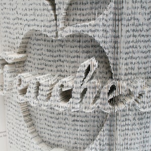 May include: A book with pages folded into a three-dimensional design, creating the word "Embrace" in a white, gray, and black color scheme.