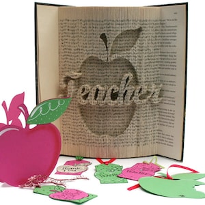 May include: A book with a red apple and the word "Teacher" cut out of the pages. The book is open and the pages are folded to create the shape of the apple. There are several small paper apple tags with the words "Thank You", "Teacher", and "Apple" on them.